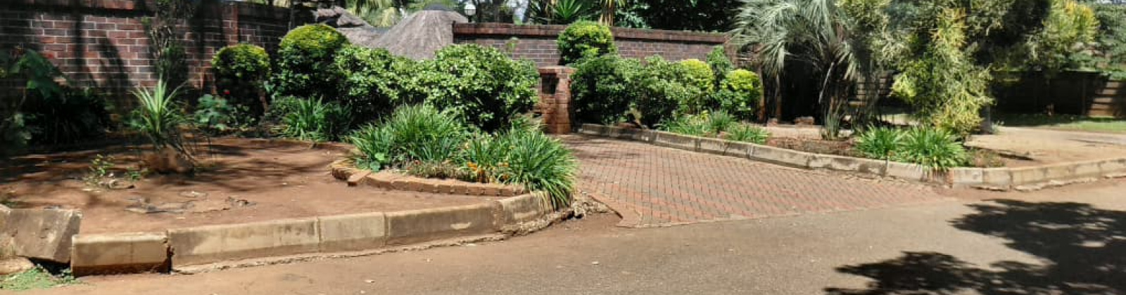 A view of the secure entrance area and landscaped boundary wall of the property. The scene features a high, red-brick perimeter wall lined with lush green bushes, ornamental shrubs, and tall palm trees under a partly cloudy sky. A brick-paved driveway slopes gently toward a gated entrance or opening in the wall. The foreground consists of an asphalt driveway or roadway with some shaded patches, bordered by a concrete curb and a partially cleared garden bed ready for planting. A view of the secure entrance area and landscaped boundary wall of the property. The scene features a high, red-brick perimeter wall lined with lush green bushes, ornamental shrubs, and tall palm trees under a partly cloudy sky. A brick-paved driveway slopes gently toward a gated entrance or opening in the wall. The foreground consists of an asphalt driveway or roadway with some shaded patches, bordered by a concrete curb and a partially cleared garden bed ready for planting.